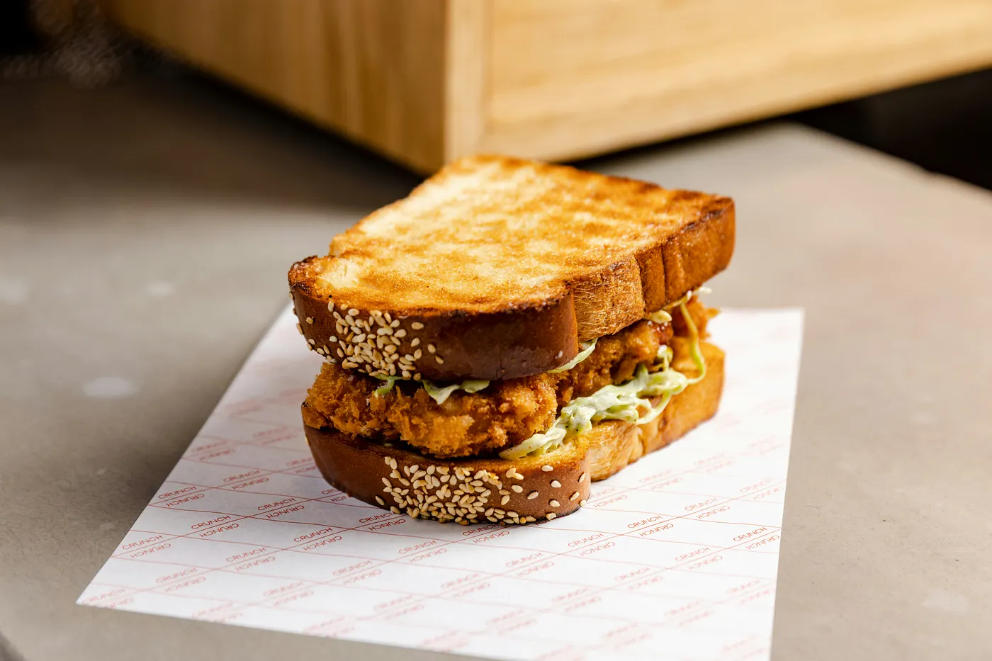 A fried chicken sandwich stacked between two thick slices of golden-toasted sesame seed milk bread, filled with a crispy breaded chicken patty and creamy coleslaw. The sandwich rests on white wax paper printed with a repeating red "CRUNCH" grid pattern, set on a gray concrete surface with a blurred wooden box in the background.